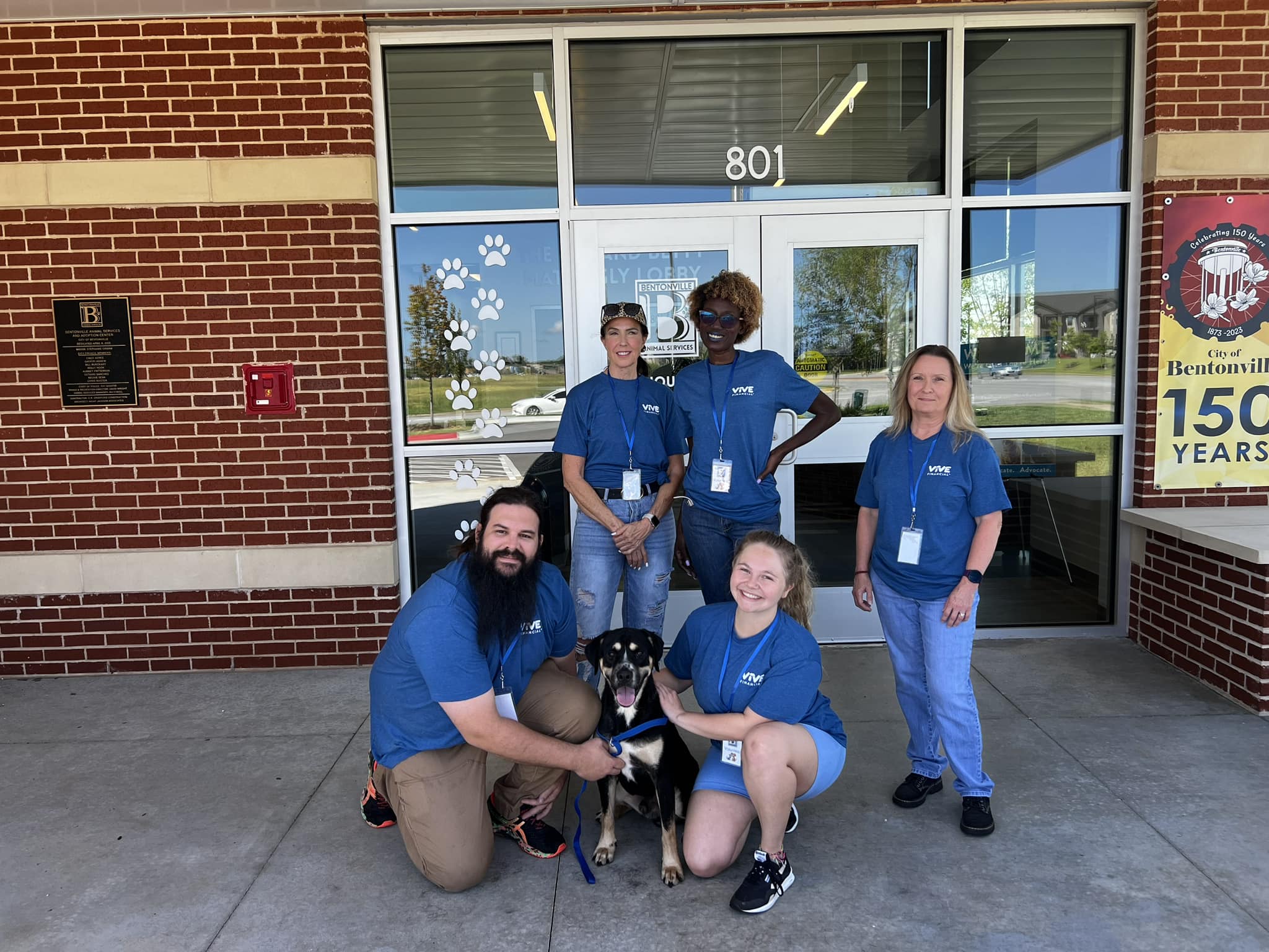 Group photo of five volunteers wearing blue shirts, posing outside the Bentonville Animal Services building with a black and tan dog.